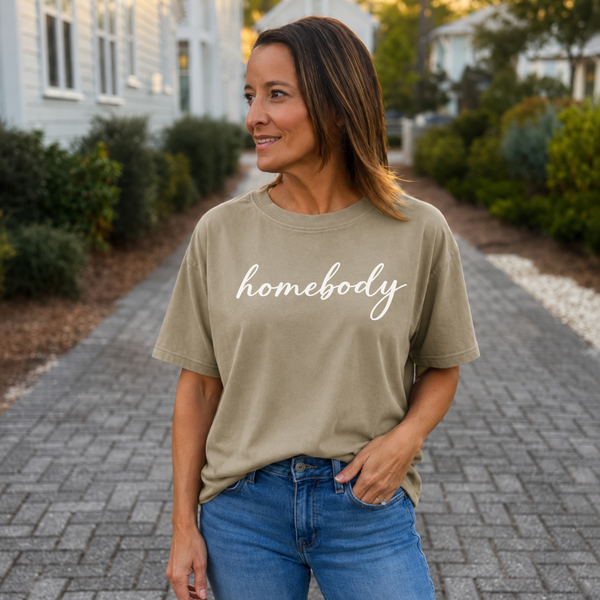 A woman standing outside with a khaki shirt on that says "homebody" in script writing