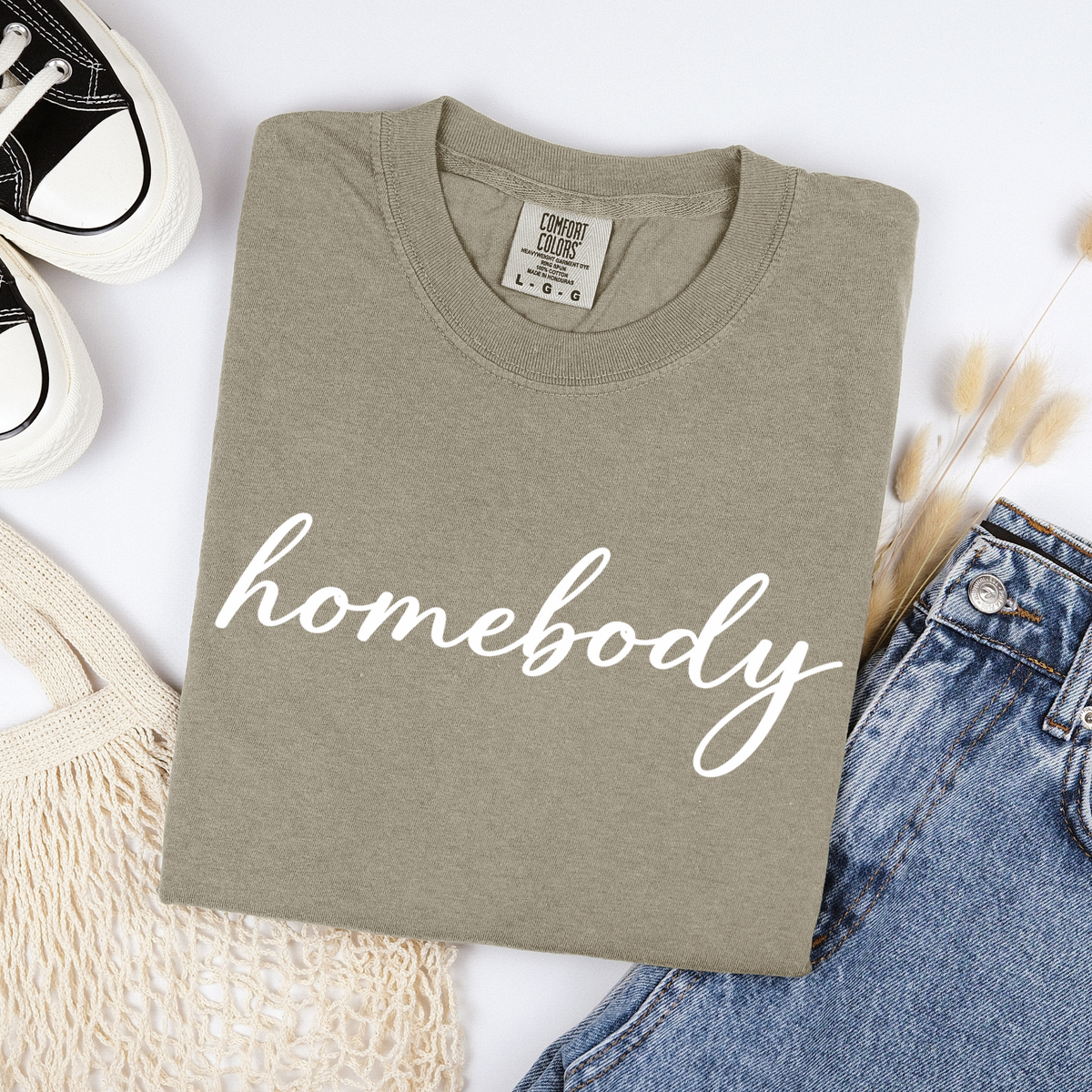 Folded khaki beige t-shirt with 'homebody' text on a white surface with jeans and sneakers.