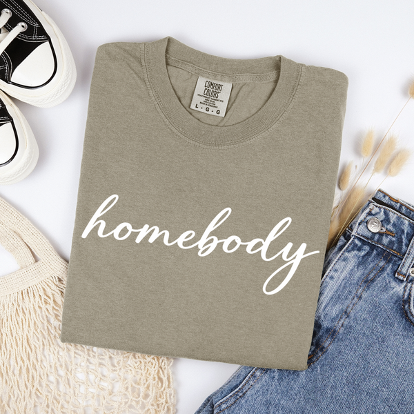Folded khaki beige t-shirt with 'homebody' text on a white surface with jeans and sneakers.