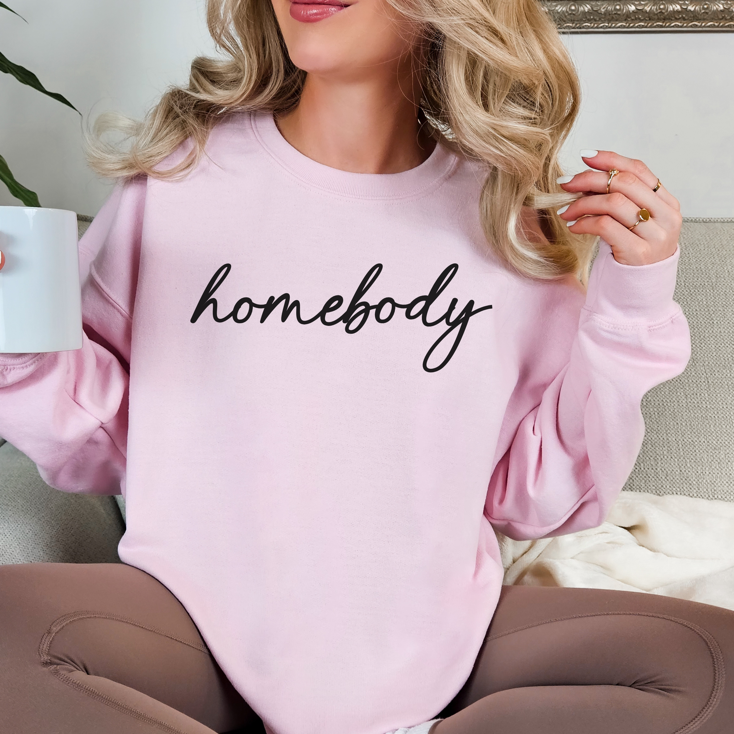 Person wearing a pink sweatshirt with 'homebody' text, holding a mug.