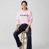 Woman wearing a pink 'homebody' sweatshirt sitting on a stool against a plain background