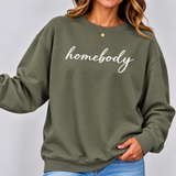 A woman wearing an olive green crewneck sweatshirt with the word 'homebody' printed in white script across the chest, paired with blue jeans against a light gray background.