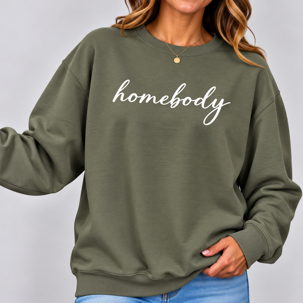 A woman wearing an olive green crewneck sweatshirt with the word 'homebody' printed in white script across the chest, paired with blue jeans against a light gray background.