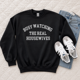 A black crewneck sweatshirt with white text reading 'BUSY WATCHING THE REAL HOUSEWIVES' is laid flat on a quilted cream bedspread, styled with light blue jeans and white sneakers.