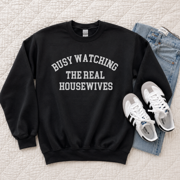 A black crewneck sweatshirt with white text reading 'BUSY WATCHING THE REAL HOUSEWIVES' is laid flat on a quilted cream bedspread, styled with light blue jeans and white sneakers.