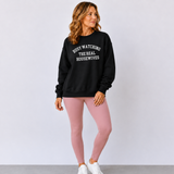 A woman modeling a black crewneck sweatshirt with white text that reads 'BUSY WATCHING THE REAL HOUSEWIVES' paired with dusty pink leggings and white sneakers, set against a plain light gray studio background.