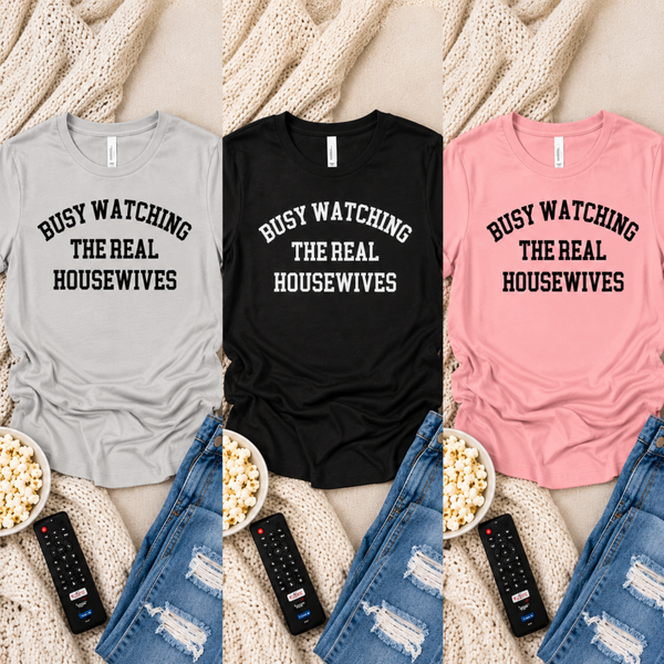 Three women's short-sleeve graphic T-shirts shown in a flat-lay collage: light gray, black, and pink. Each shirt has the phrase 'BUSY WATCHING THE REAL HOUSEWIVES' printed across the chest in arched lettering.