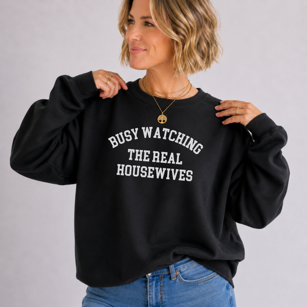 A woman wearing a black crewneck sweatshirt with white text that reads 'BUSY WATCHING THE REAL HOUSEWIVES', paired with blue jeans, against a plain light gray background.