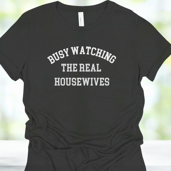 Black t-shirt with 'BUSY WATCHING THE REAL HOUSEWIVES' text on a blurred background