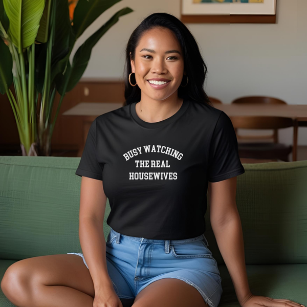 Woman sitting on couch wearing a black Tee with white lettering that reads "Busy Watching the Real Housewives."