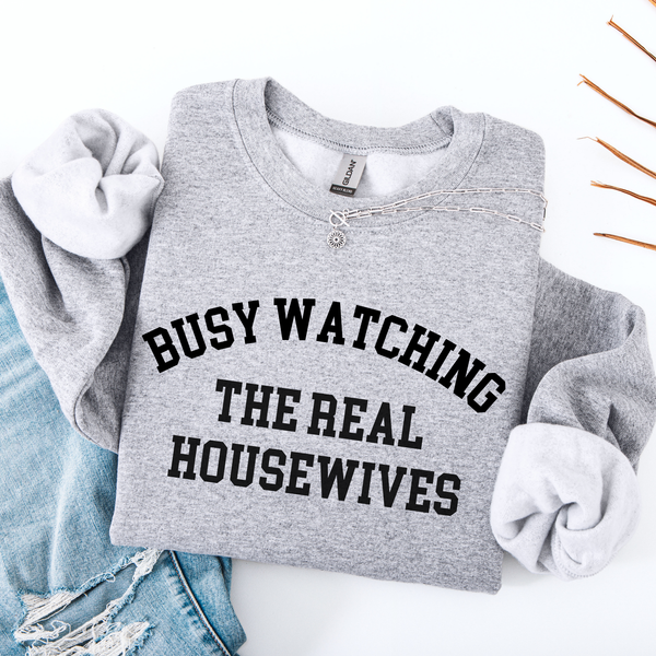 Gray folded sweatshirt with 'Busy Watching The Real Housewives' text on a white background