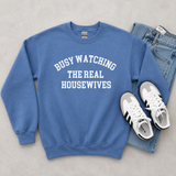 A blue crewneck sweatshirt with white text reading 'BUSY WATCHING THE REAL HOUSEWIVES' is laid flat on a light gray surface, styled with blue jeans and white sneakers.