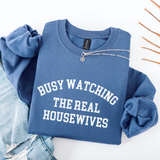 Blue folded sweatshirt with 'BUSY WATCHING THE REAL HOUSEWIVES' text on a white background