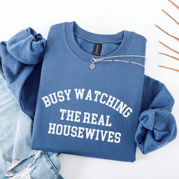 Blue folded sweatshirt with 'BUSY WATCHING THE REAL HOUSEWIVES' text on a white background