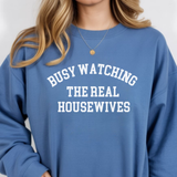 A woman wearing a blue sweatshirt with text 'BUSY WATCHING THE REAL HOUSEWIVES' on a plain background