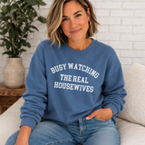 A woman wearing a dusty blue crewneck sweatshirt with the text 'BUSY WATCHING THE REAL HOUSEWIVES' in white arched lettering, sitting on a light beige couch in front of a white brick wall.