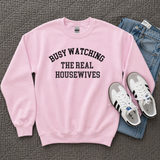 A pink crewneck sweatshirt with black text reading 'BUSY WATCHING THE REAL HOUSEWIVES' is laid flat on a gray quilted blanket, styled with blue jeans and white sneakers.