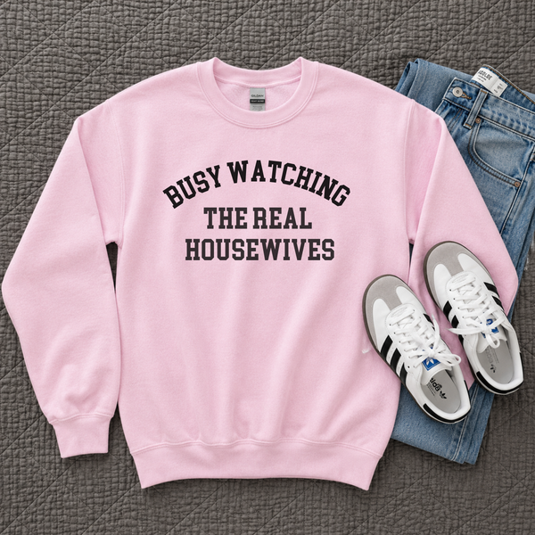 A pink crewneck sweatshirt with black text reading 'BUSY WATCHING THE REAL HOUSEWIVES' is laid flat on a gray quilted blanket, styled with blue jeans and white sneakers.