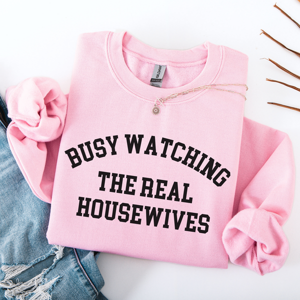 Pink folded sweatshirt with 'BUSY WATCHING THE REAL HOUSEWIVES' text on a white background