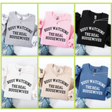 Collage of six crewneck sweatshirts in different colors (Gray, Pink, Black, White, Sand, Blue) with 'BUSY WATCHING THE REAL HOUSEWIVES' text.