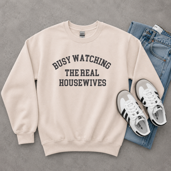 A cream-colored crewneck sweatshirt with the text 'BUSY WATCHING THE REAL HOUSEWIVES' printed in black arched lettering on the chest. The sweatshirt is styled in a flat lay with blue jeans and white and black sneakers on a gray textured background.