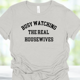 Gray t-shirt with 'BUSY WATCHING THE REAL HOUSEWIVES' text on a blurred background