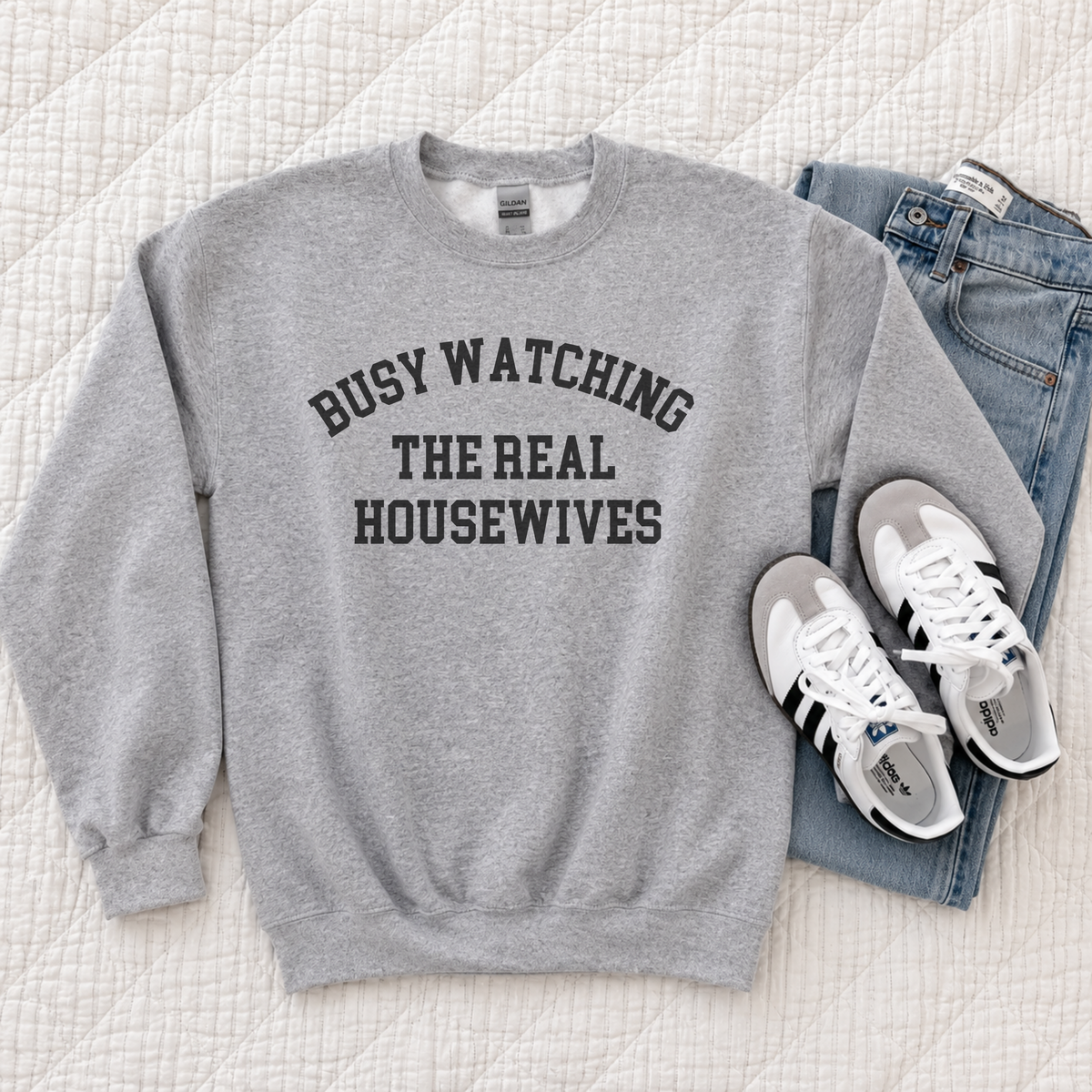 A gray sweatshirt with the words "Busy Watching the Real Housewives" on the front laying on top of a pair of jeans along side a pair of sneakers.