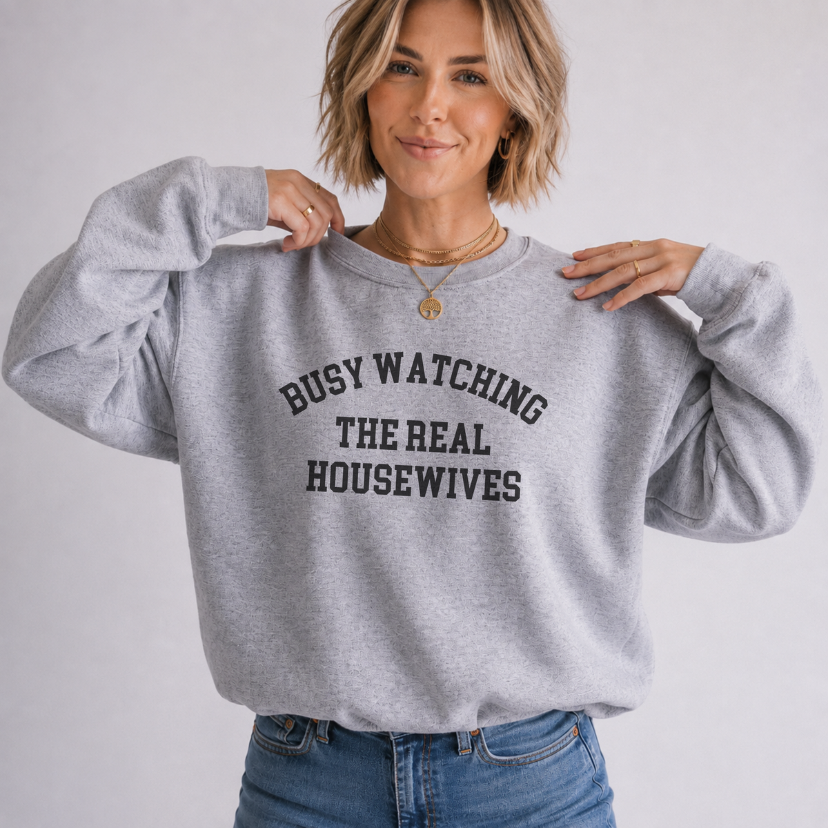 A woman wearing a light heather gray crewneck sweatshirt with the text 'BUSY WATCHING THE REAL HOUSEWIVES' printed in black arched letters across the chest, paired with blue jeans against a plain light gray background.