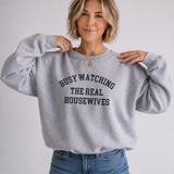 A woman wearing a light heather gray crewneck sweatshirt with the text 'BUSY WATCHING THE REAL HOUSEWIVES' printed in black arched letters across the chest, paired with blue jeans against a plain light gray background.