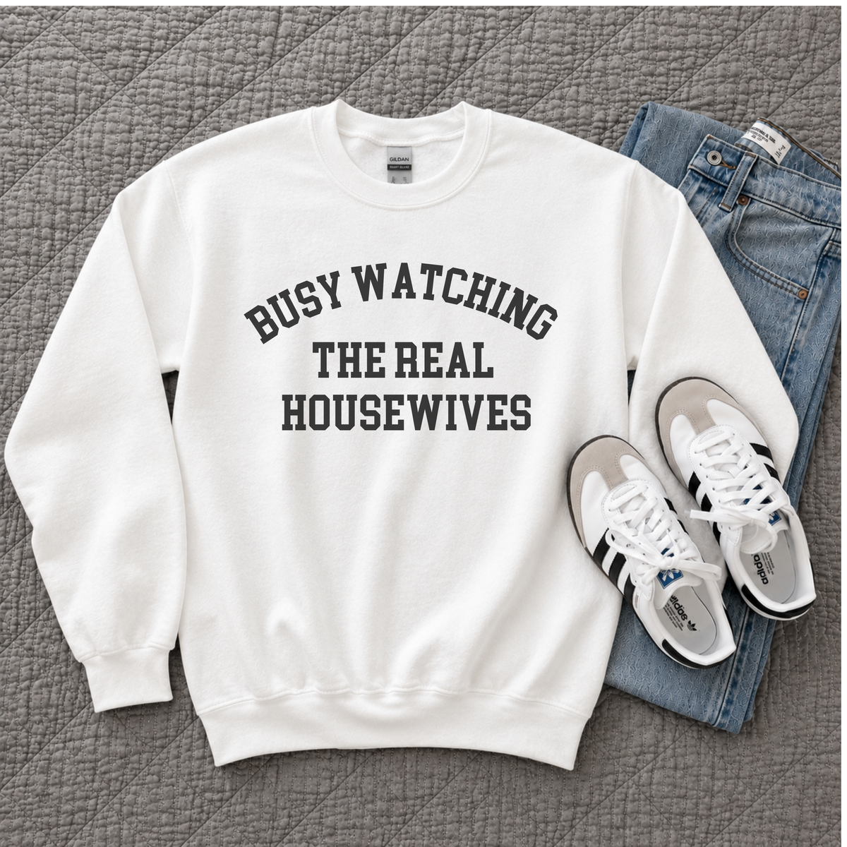 A white crewneck sweatshirt with the text 'BUSY WATCHING THE REAL HOUSEWIVES' printed in black arched letters on the chest, styled in a flat lay with blue jeans and white sneakers on a gray quilted blanket.