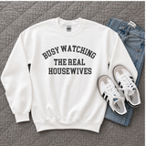 A white crewneck sweatshirt with the text 'BUSY WATCHING THE REAL HOUSEWIVES' printed in black arched letters on the chest, styled in a flat lay with blue jeans and white sneakers on a gray quilted blanket.
