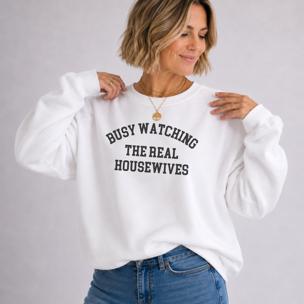 A woman wearing a white crewneck sweatshirt with black text that reads 'BUSY WATCHING THE REAL HOUSEWIVES', paired with blue jeans, against a light gray studio background.