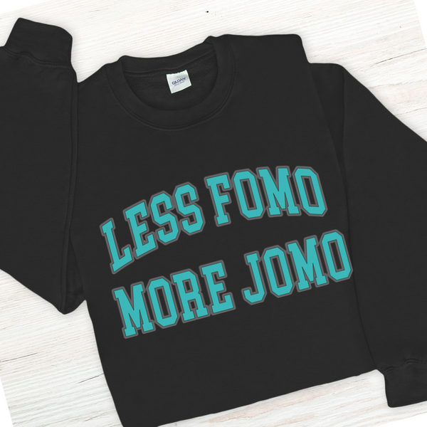 Black sweatshirt with teal text 'LESS FOMO MORE JOMO' on a white wooden surface