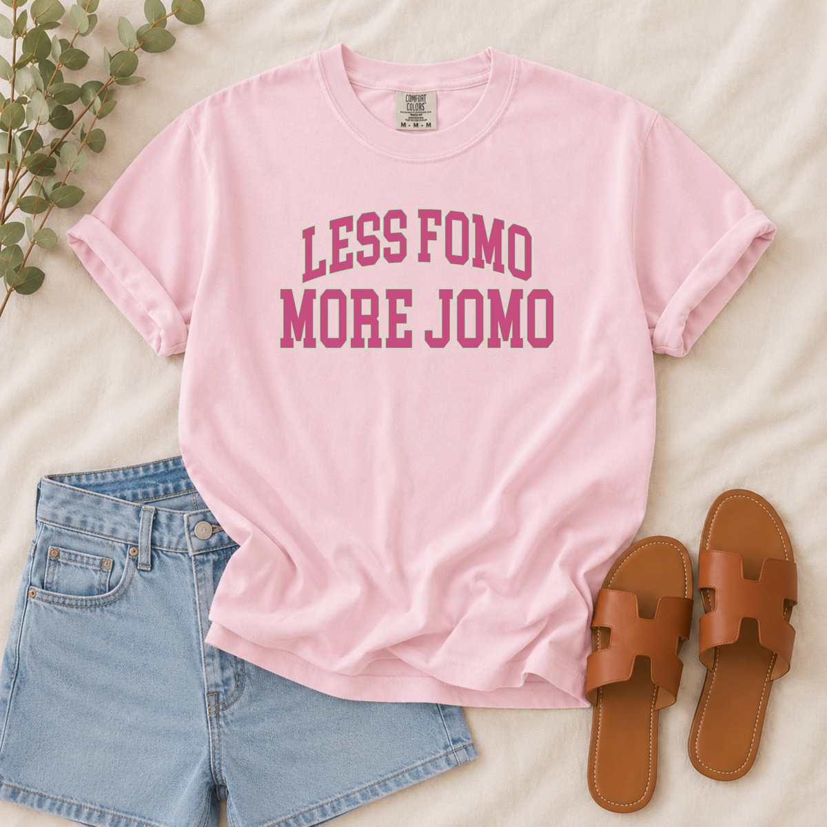A light pink short-sleeve graphic T-shirt laid flat on a cream fabric background, styled with light blue denim shorts, brown slide sandals, and a sprig of green leaves. The shirt has rolled sleeves and the text 'LESS FOMO MORE JOMO' printed in pink across the chest.