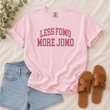 A light pink short-sleeve graphic T-shirt laid flat on a cream fabric background, styled with light blue denim shorts, brown slide sandals, and a sprig of green leaves. The shirt has rolled sleeves and the text 'LESS FOMO MORE JOMO' printed in pink across the chest.
