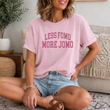 A woman sitting cross-legged on a patterned rug in a living room setting, wearing a light pink t-shirt with the text 'LESS FOMO MORE JOMO' printed in pink block letters across the chest, paired with denim shorts and brown sandals.