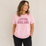 A woman wearing a light pink crewneck t-shirt with the text 'LESS FOMO MORE JOMO' printed in dark pink across the chest, paired with black leggings, set against a plain light beige background.