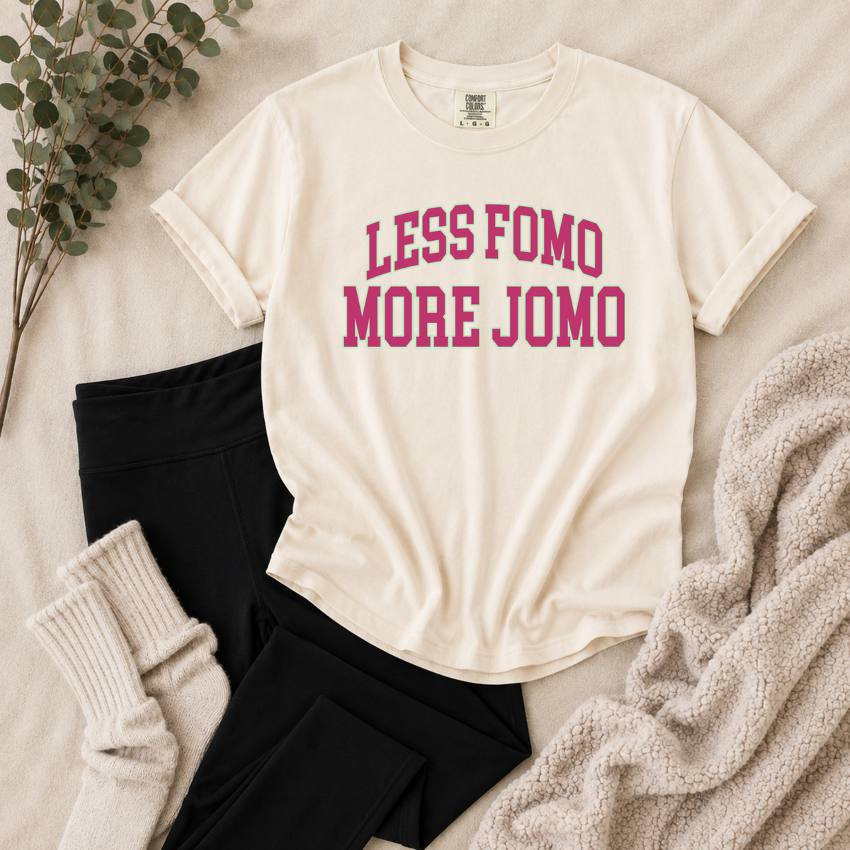 An off-white short-sleeve graphic T-shirt with the text 'LESS FOMO MORE JOMO' printed in pink across the chest, styled in a flat lay with black leggings, cream knit socks, a chunky knit blanket, and eucalyptus branches on a light fabric background.