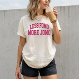 A woman wearing a cream-colored t-shirt with pink text that reads 'LESS FOMO MORE JOMO' in a casual outdoor setting, holding an iced coffee.
