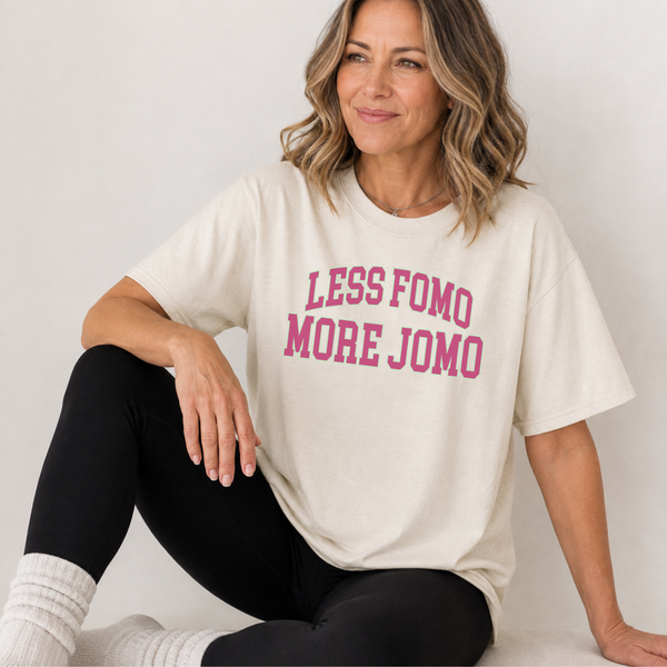A woman sitting and wearing an oversized cream-colored t-shirt with the text 'LESS FOMO MORE JOMO' printed in pink across the chest, paired with black leggings and white socks.