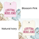 Two folded graphic T-shirts shown in a collage: one light pink labeled 'Blossom Pink' and one cream/off-white labeled 'Natural Ivory'. Both have the slogan 'LESS FOMO MORE JOMO' in pink lettering.