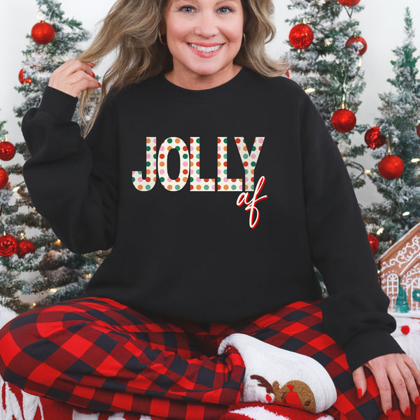 Person wearing a black crewneck with 'Jolly AF' text in festive colors, sitting in front of Christmas trees.