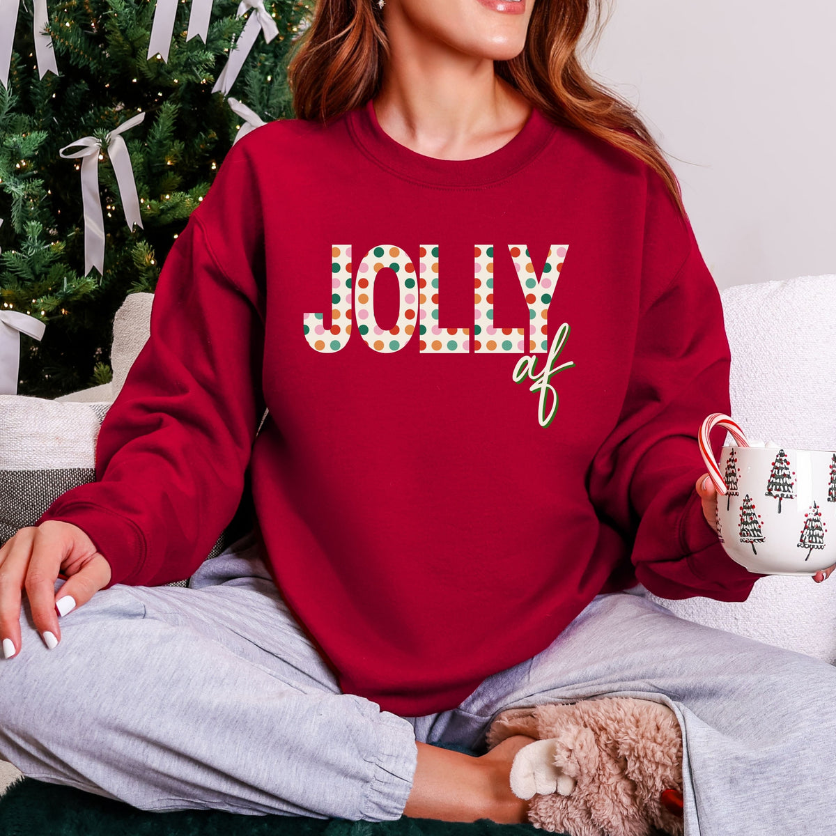 Woman wearing a red sweatshirt with Jolly AF text, sitting in front of a Christmas tree.