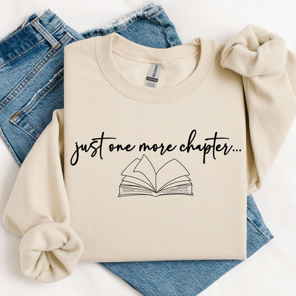 Beige sweatshirt with 'just one more chapter...' text and book graphic on a white background with blue jeans.