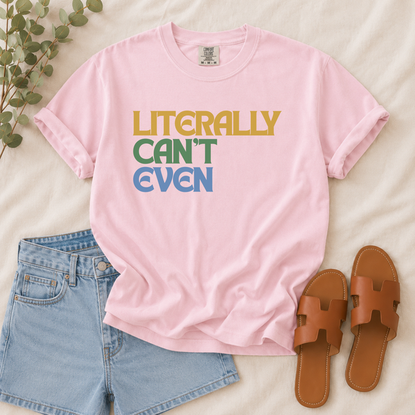 A light pink graphic t-shirt with the text 'LITERALLY CAN'T EVEN' in yellow, green, and blue block letters, styled in a flat lay with light blue denim shorts, brown slide sandals, and a sprig of green eucalyptus on a beige fabric background.