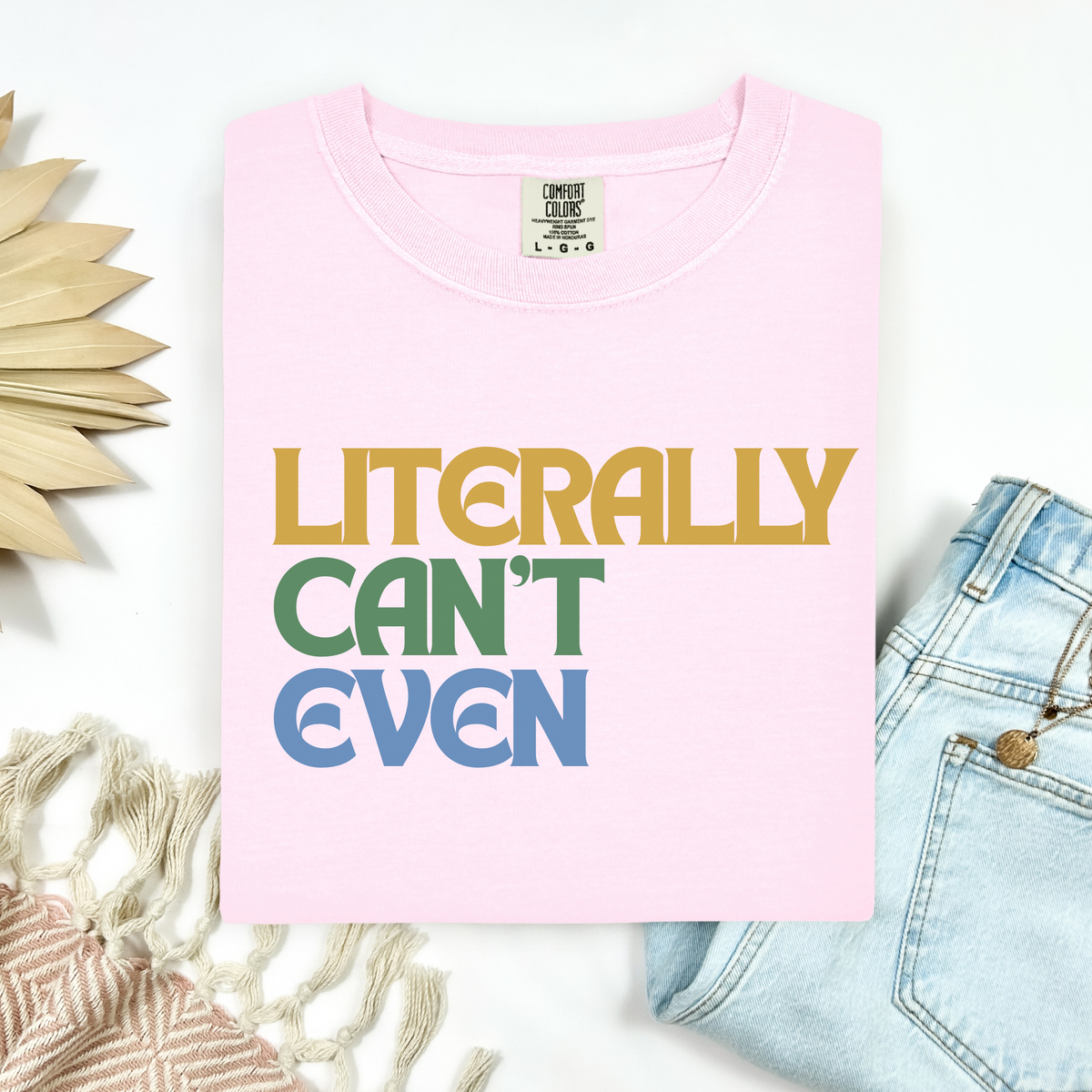 A folded light pink t-shirt with the text 'LITERALLY CAN'T EVEN' printed in yellow, green, and blue block letters. The shirt is styled in a flat-lay arrangement with light blue jeans, a beige woven textile, and a decorative wooden leaf on a white background.