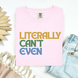 A folded light pink t-shirt with the text 'LITERALLY CAN'T EVEN' printed in yellow, green, and blue block letters. The shirt is styled in a flat-lay arrangement with light blue jeans, a beige woven textile, and a decorative wooden leaf on a white background.