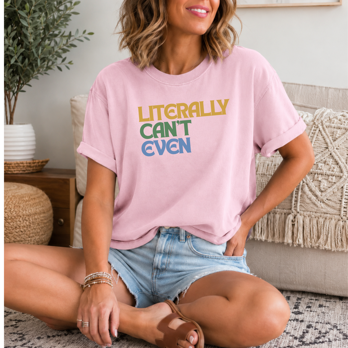 A woman sitting cross-legged in a living room wearing a light pink graphic t-shirt with the text 'LITERALLY CAN'T EVEN' in yellow, green, and blue. She is wearing denim shorts and brown slide sandals.