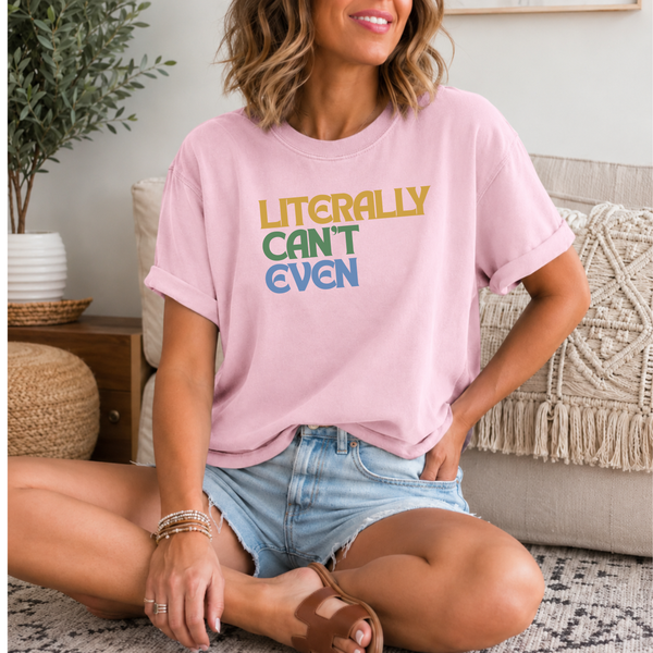 A woman sitting cross-legged in a living room wearing a light pink graphic t-shirt with the text 'LITERALLY CAN'T EVEN' in yellow, green, and blue. She is wearing denim shorts and brown slide sandals.