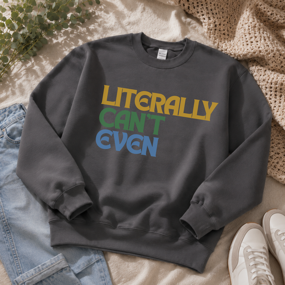 A dark gray crewneck sweatshirt with the text 'LITERALLY CAN'T EVEN' printed in yellow, green, and blue retro lettering. The sweatshirt is styled in a flat-lay arrangement on a beige blanket with light blue jeans, a knit throw blanket, and white sneakers.